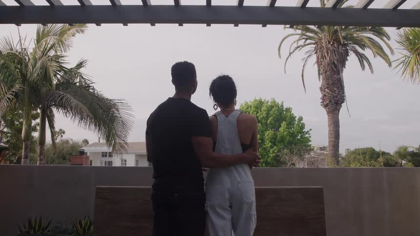 A couple stands with their backs to the camera looking out over a suburban landscape with palm trees framed by a dark pergola structure the man has his arm around the woman who is wearing lightcolored overalls suggesting a moment of shared contempla