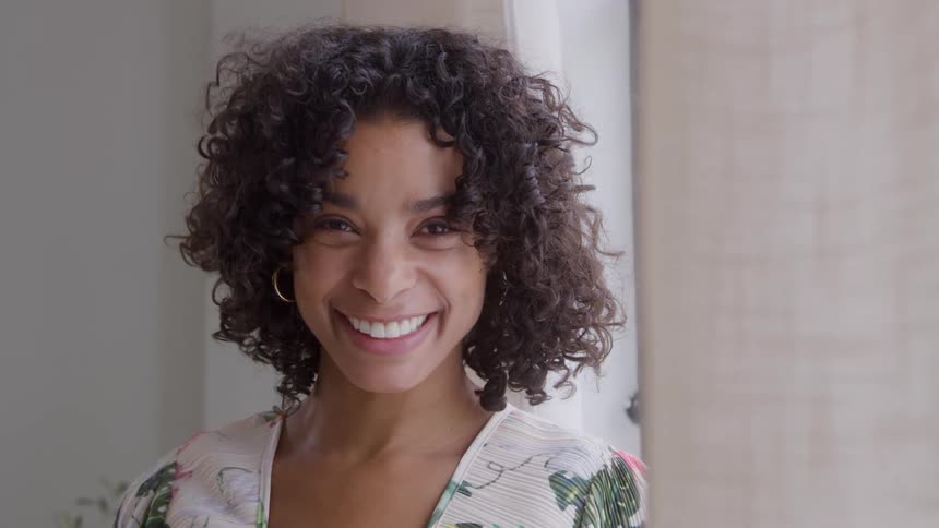 The video is a closeup of a young woman with dark curly hair and hoop earringsshe is smiling widely and genuinely at the camera standing by a window with natural light filtering through sheer curtains