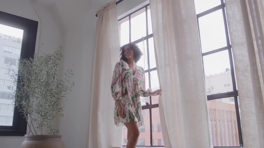 A joyful mixedrace woman in a tropical dress is looking forward next to tall windows with sheer curtains