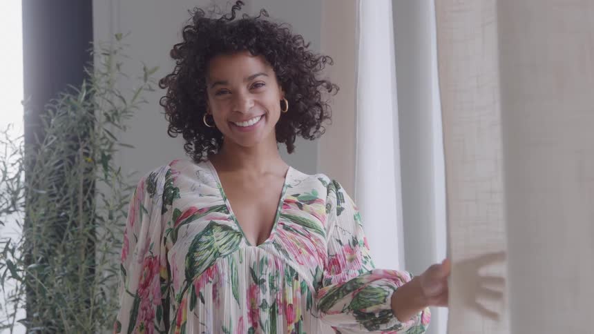 A young woman in a bright summer dress stands beside a curtain holding it gently with a flower plant behind her as she smiles happily
