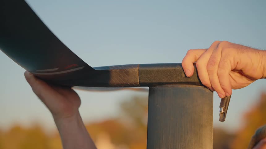 This closeup video shows a pair of hands assembling or adjusting a piece of outdoor equipment likely securing a black curved component to a metallic post the hands are working with a small metal pin or tool against a background of soft golden light