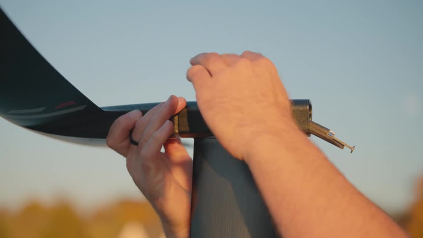 This closeup video shows a persons hands securing a dark curved piece of material possibly a flexible blade or vane into a metallic connection piece atop a vertical post the scene is outdoors with a bright sky and warm diffused background light su