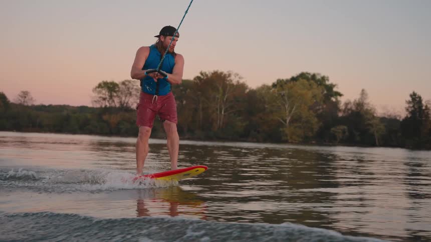A man wake surfs on a lake with trees in the background at sunset