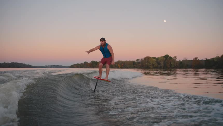 A man on an electric hydrofoil board wake surfs on a lake during sunset skimming the water and riding a wave behind a boat