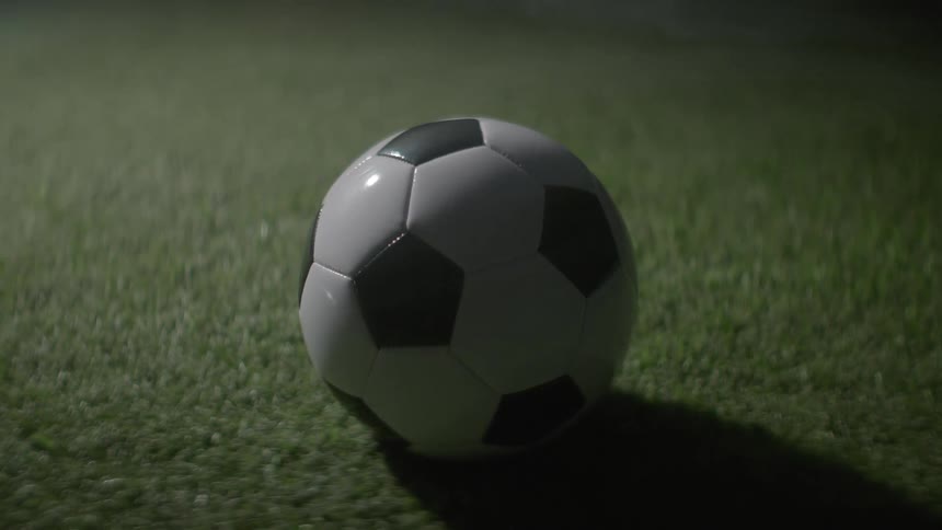 A close view of a black and white soccer ball resting on green grass illuminated by a low light source that casts a deep shadow