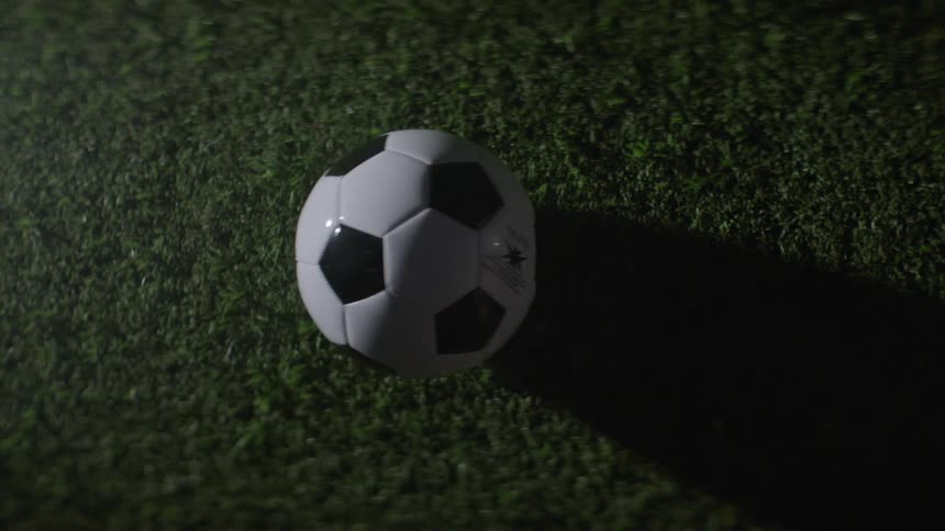 A closeup overhead shot shows a classic black and white panel soccer ball resting static on a field of green grass turf with a harsh directional light source casting a long dark shadow of the ball across the ground