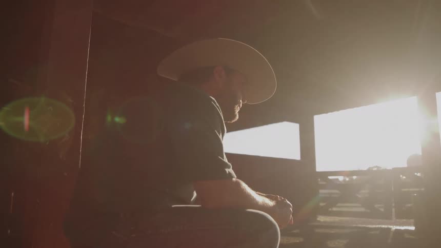 A man wearing a widebrimmed cowboy hat and dark clothing is seen in profile seated in the darkness of a barn looking out toward an intensely bright opening