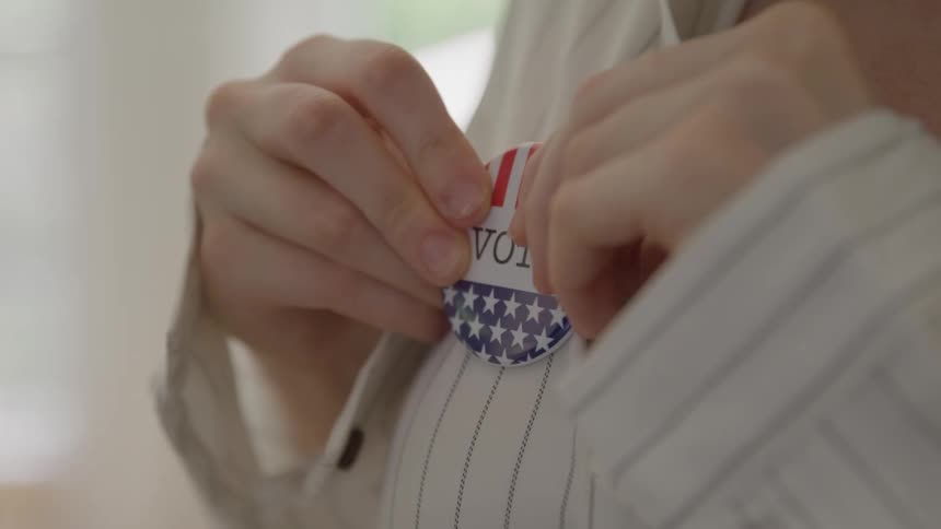 This is a closeup video showing a persons hands pinning an i voted sticker or button onto the front of their striped shirtthe ovalshaped button features a design based on the american flag signifying participation in an election