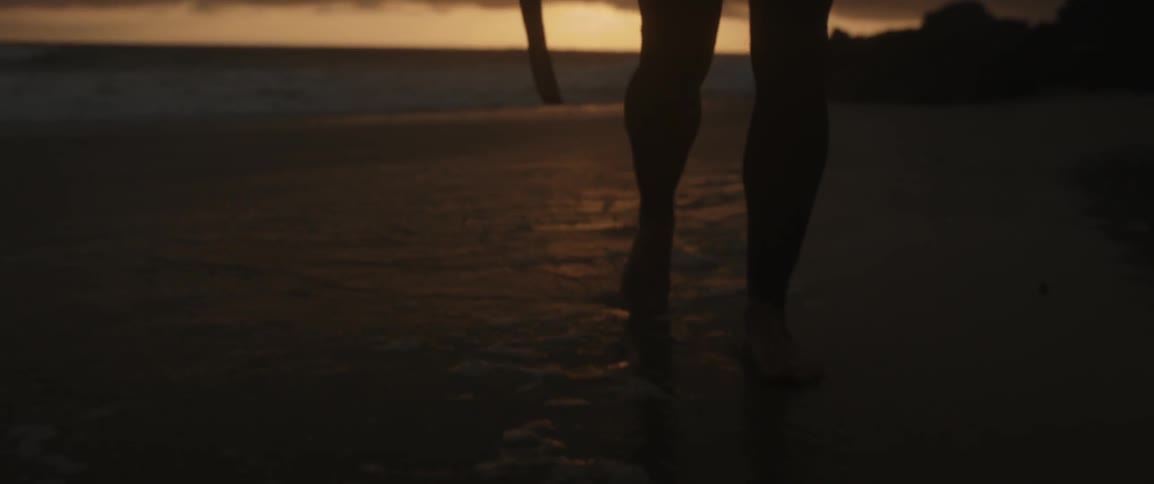 A surfer walks on a sandy beach at sunset