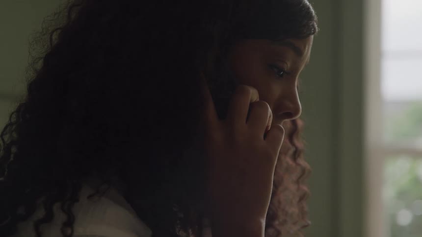 A young black woman sits indoors at midday holding a smartphone to her ear as she answers a call looking slightly sad in the room
