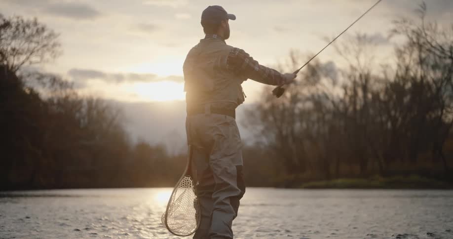 A man wearing waders and a cap is fly fishing in a shallow river at sunset