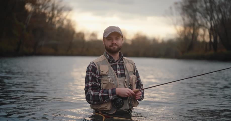 A young man stands in the river at midday wearing a hat holding a fishing line as he fishes peacefully