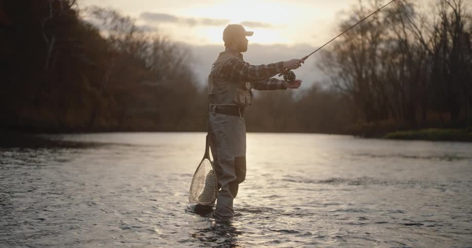 A person in fishing waders is fly fishing while standing in a shallow river at sunset