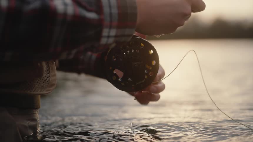 A person wearing a plaid shirt and waders flyfishing holding a reel dripping with water in soft light