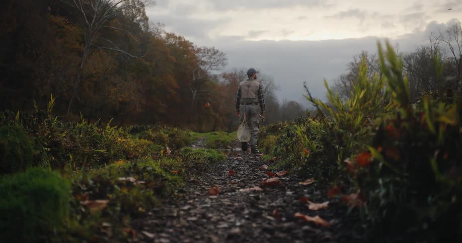 A lowangle shot follows a person walking away down a leafstrewn dirt path surrounded by lush green and autumnal foliage the person is dressed in outdoor gear and appears to be carrying a fishing net as they head towards a misty valley under a cloudy