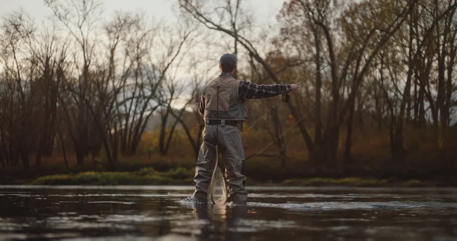 A man wearing waders fly fishes in a river surrounded by bare trees at sunset
