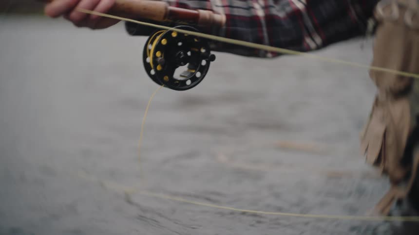 A close view of a persons hands holding a fly fishing rod with a reel letting out a bright yellow fishing line over rippling water
