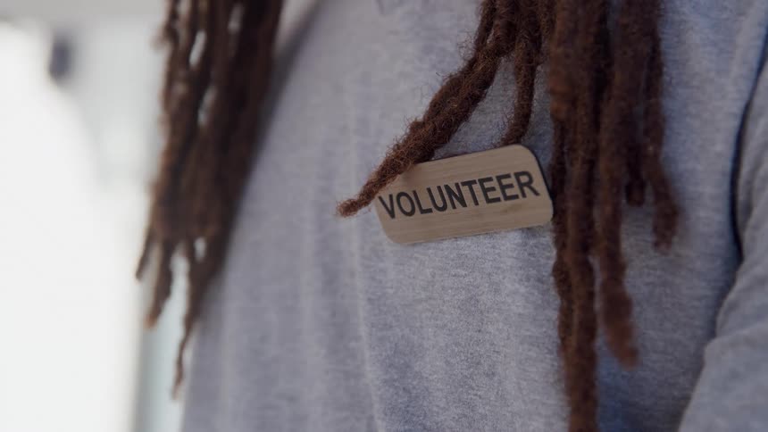 A man in a grey tshirt with long dreadlocks is wearing a prominent golden tag with the word volunteer in a heavy black font
