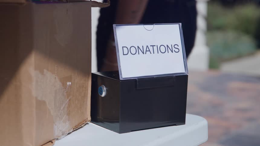 A black donation box with a donations sign in a plastic holder is sitting on a white table next to a cardboard box and a woman passes by the side wearing jeans
