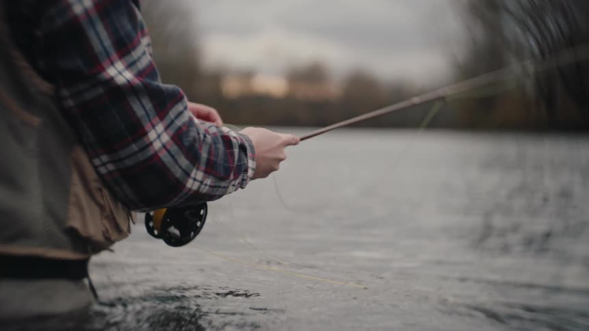 A closeup shot of a person standing waistdeep in moving water wearing a plaid shirt and fishing waders actively casting a fly fishing line with repeated forward and backward motions of the rod and reel