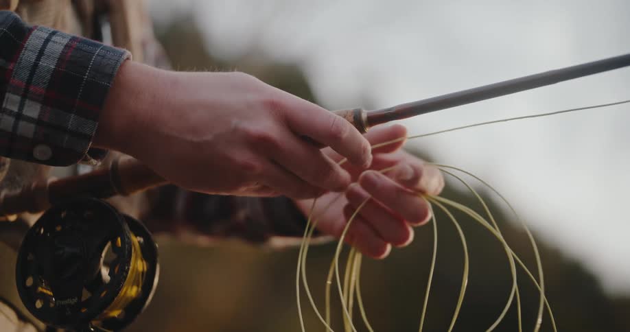 Hands adjust fishing line with fly rod and yellow reel in softly blurred outdoor setting