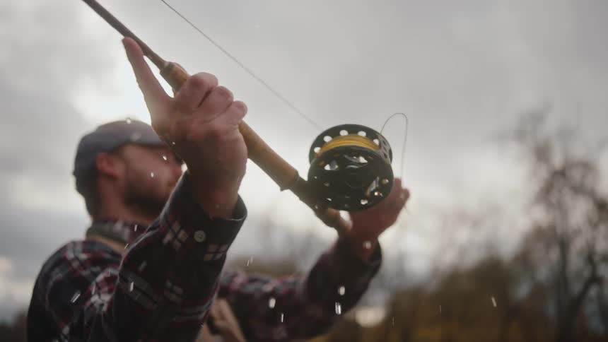 A bearded man in a plaid shirt and cap raises a fishing rod with water dripping from the reel against a bright cloudy sky