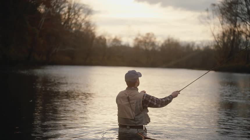 Person fly fishing in calm water reflecting golden light among leafless trees