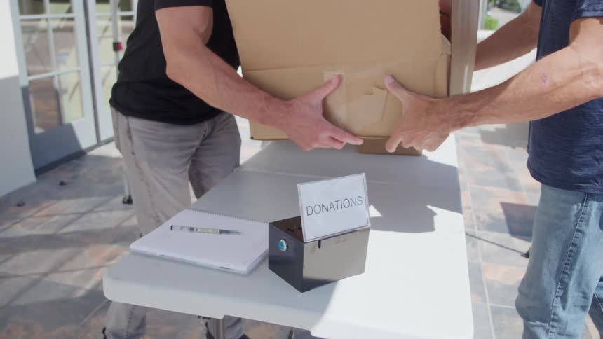 Two people lifting a large cardboard box over a white folding table with a donation box