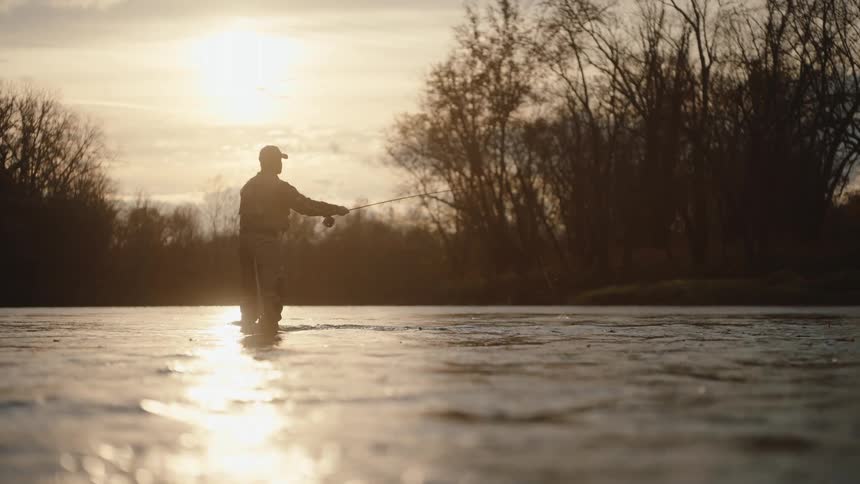 Silhouette of a man wearing a baseball cap fishing with his knees underwater outdoors during dusk or dawn
