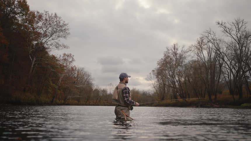 A man stands in the river on a cloudy day swinging his fishing line as he fishes with leafless trees lining both sides of the river