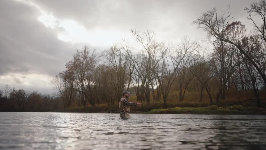 A fisherman wearing waders stands in a wide river on a cloudy overcast day fly fishing with a long rodthe water is calm and reflects the low light with a dense wooded bank of mostly bare trees visible in the background