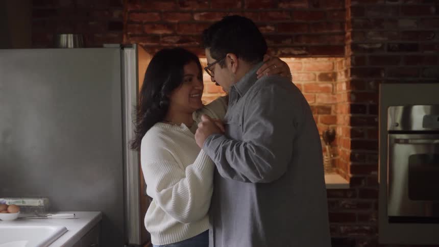 A young woman is washing dishes at the kitchen sink when her husband walks in holds her and they begin dancing together in the kitchen