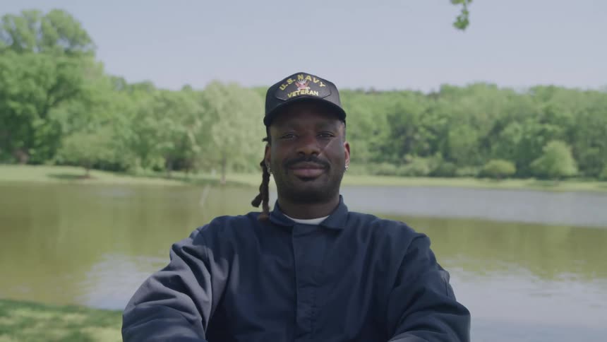 A man wearing a dark blue jumpsuit and a us navy veteran baseball cap sitting outdoors with a calm expression with a lake and lush green trees forming the background
