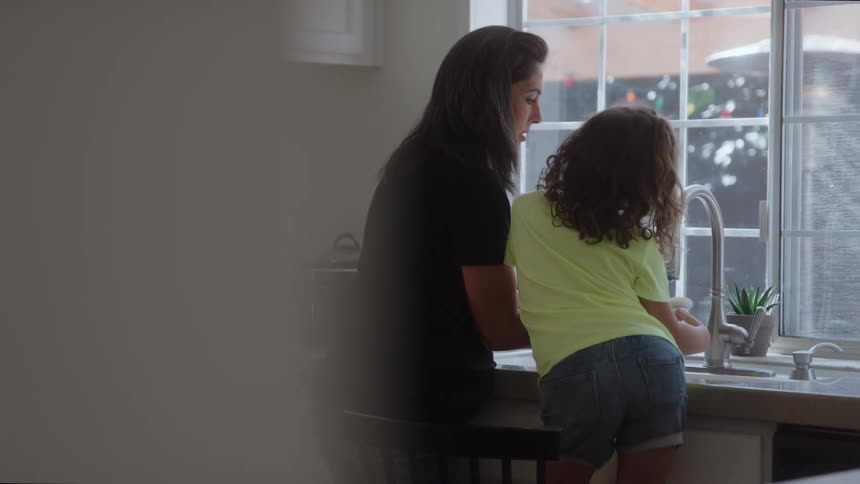 Adult and child together at kitchen sink lit by window with backyard view