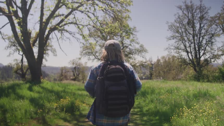 A person wearing a plaid shirt a baseball cap and a black backpack is seen walking away from the camera down a grassy paththey are in a sunny outdoor setting surrounded by trees and wildflowers seemingly on a hike or nature walk
