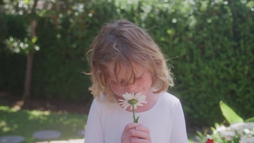 A child stands in the garden at midday holding a flower gently to her nose as she smells it causing her to smile