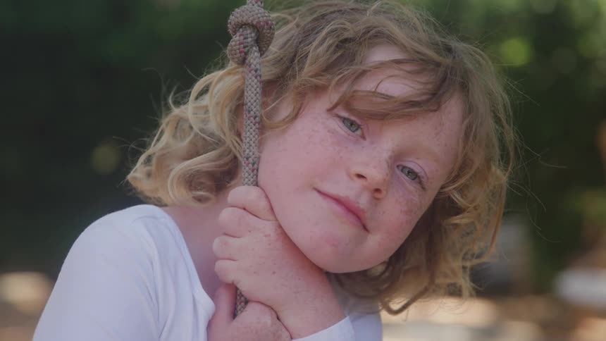 A young child with blonde curly hair and freckles holds onto a knotted rope looking forward with a slight smile