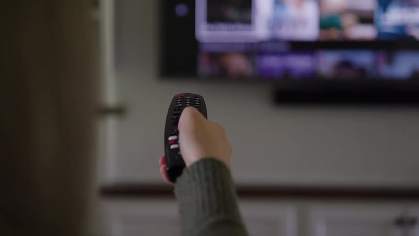 A closeup of a hand operating a television with a remote aimed at a glowing blurred screen