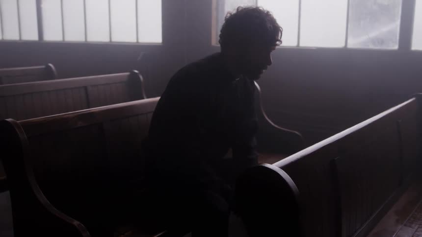 A dark silhouette of a man sits alone in a church pew looking down in a dimly lit somber interior with bright windows in the background