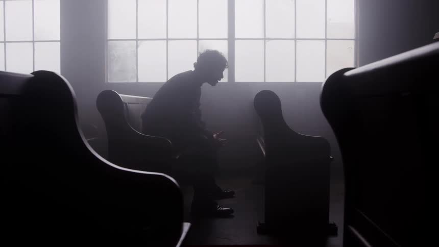 Silhouette of a young man praying while sitting on a pew in a church during daytime