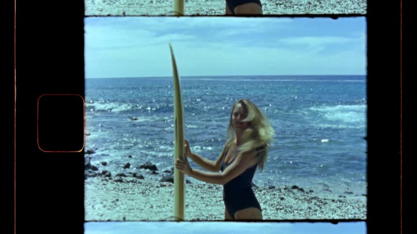 A young woman in a black swimsuit with long blonde hair holds a yellow surfboard and smiles at the camera on a rocky beach by the sparkling ocean