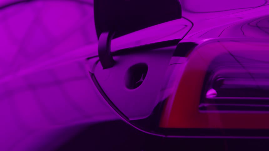 A hand is plugging a charger into the port of a dark vehicle bathed in striking magenta and purple light