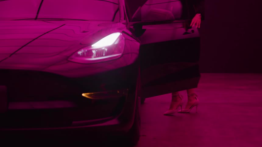 A black sedan with its bright headlights on illuminated by intense magenta and pink ambient lighting as a woman in a short black dress and high heels walks toward the car reaches for the handle and opens the door to enter