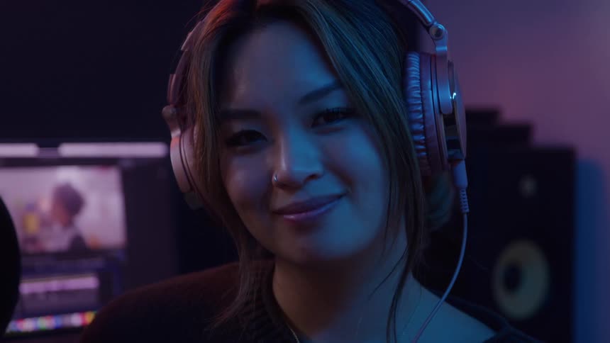 A smiling young woman wearing pink headphones looks directly at the camera in a dark room illuminated by blue and pink ambient light with audio equipment visible