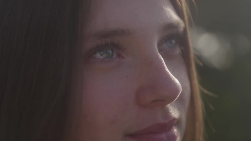 An extreme close side view of a young womans face with bright blue eyes looking upwards in soft sunlight outdoors
