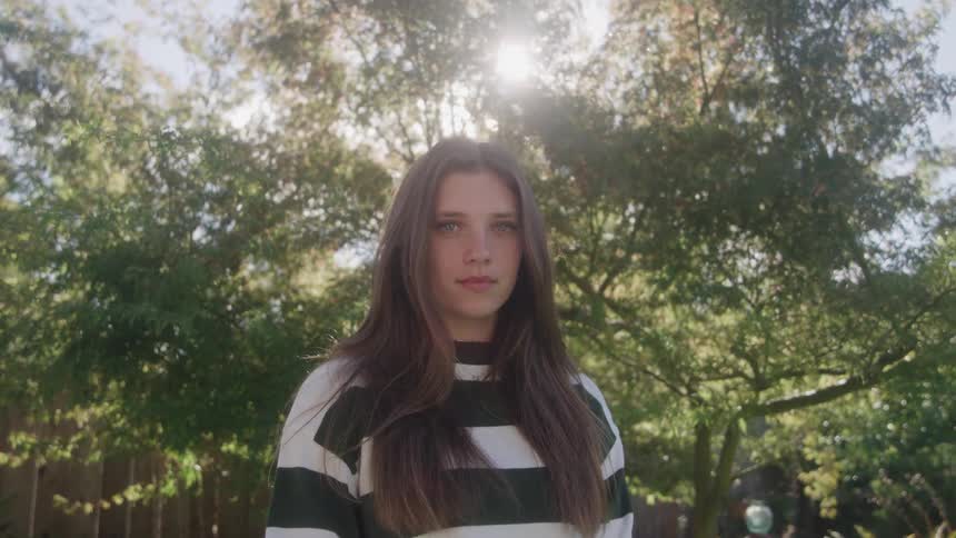 A medium static shot features a young woman with long dark hair wearing a striped sweater looking directly at the camera with a neutral expression she is outdoors backlit by the sun shining through the dense bright green leaves of trees in the backg