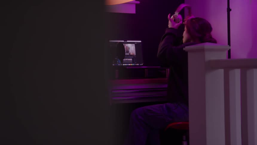 A person walks into a dimly lit room with purple ambient lighting sits down at a desk with a laptop and puts on headphones to work