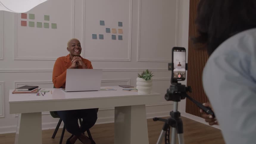A smiling african american businesswoman sits at a white desk with a laptop being recorded by a phone mounted on a tripod held by a colleague