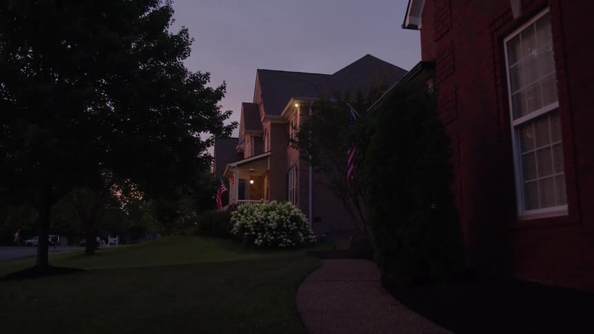 A dark exterior shot shows a quiet suburban street and large brick houses with lit porch lights along a curved concrete walkway at twilight