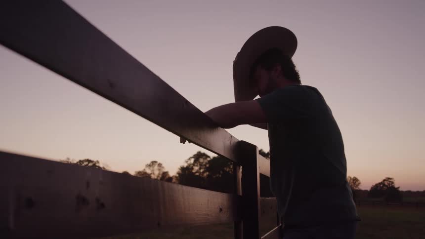 A cowboy leans against a wooden fence silhouetted against the colorful sunset sky as he holds his hat to his forehead